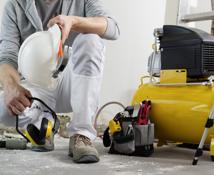 A man kneels on one knee holding a pair of noise canceling ear muffs and a hard hat. He is kneeling next to an air compressor. - air compressor filter | Choosing the Perfect Air Compressor Filter
