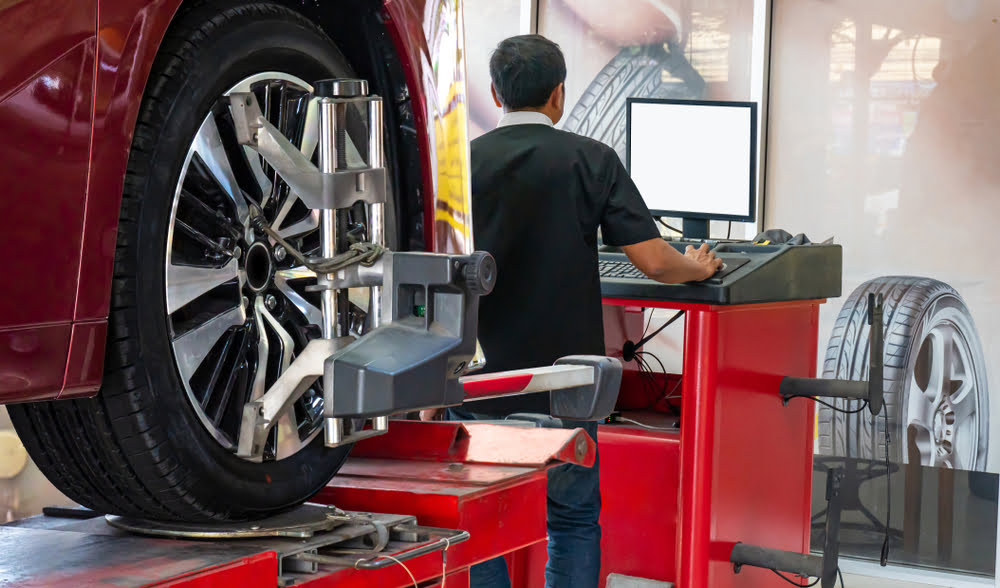 a mechanic sits in front of a computer with a tire alignment machine behind him, showing the difference between tire balancing vs alignment